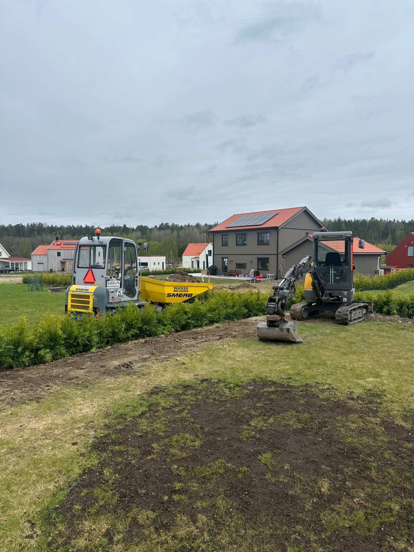 Grävmaskin och dumper utför markarbete på villatomt vid nybyggda hus